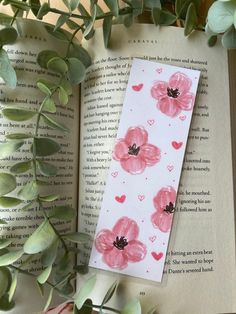 an open book with pink flowers and hearts on it next to some green leaves in the foreground