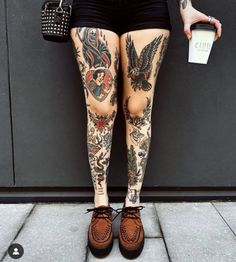 a person with tattoos on their legs holding a coffee cup and wearing brown shoes while standing in front of a wall