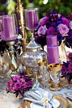 a table topped with lots of purple flowers next to gold plates and silver candlesticks