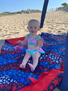 a baby sitting on top of a blue and red blanket next to the ocean under an umbrella