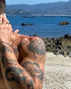 a man sitting on the beach with his hands to his face and tattoos on his chest