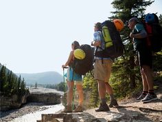 three people with backpacks are standing at the edge of a cliff overlooking a river