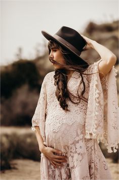 a pregnant woman wearing a black hat and white lace dress with her hands on her head