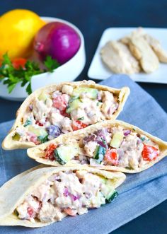 two pita breads filled with chicken salad on top of a blue napkin next to bowls of vegetables