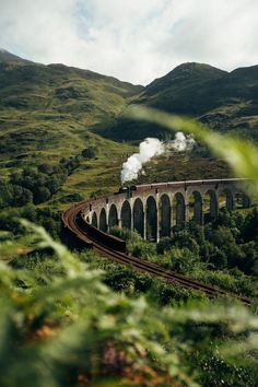 a train traveling over a bridge on top of a lush green hillside next to mountains