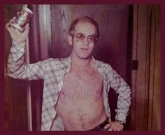 a shirtless man holding up a can of beer in his right hand and looking at the camera