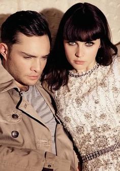 a man and woman sitting next to each other in front of a white wall wearing trench coats