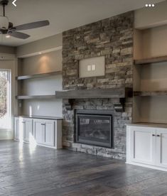 an empty living room with stone fireplace and built - in shelving units, windows, and ceiling fan