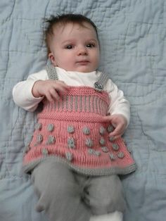 a baby laying on top of a bed wearing a pink and gray dress with pom poms