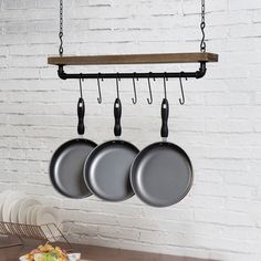 three pots and pans hanging from hooks on a white brick wall