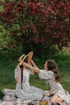 two women in dresses and hats are sitting on a blanket near a tree with pink flowers