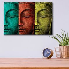 three buddha heads in different colors on a wall above a table with a clock and potted plant