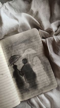 an open notebook sitting on top of a bed next to a drawing of two people holding umbrellas