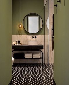 a bathroom with green walls and black and white tile flooring, along with a round mirror on the wall