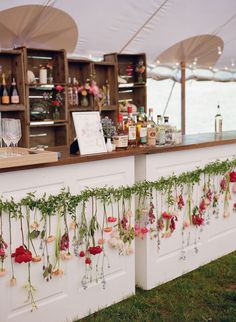 an outdoor bar with flowers hanging from the wall and wine glasses on the shelves behind it