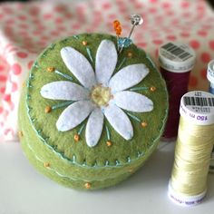 a green pillow with white flowers on it next to thread and spools