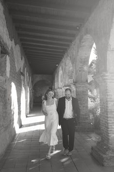 a man and woman walking down a walkway in an old brick building with sunlight coming through the windows