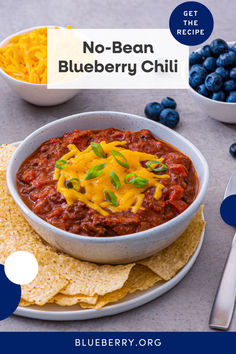 a bowl of chili with cheese and blueberries on the side, next to tortilla chips