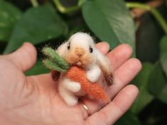 a hand holding a tiny stuffed animal with a carrot on it's back and green leaves in the background