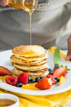 pancakes with syrup being poured over them on a white plate surrounded by berries and strawberries