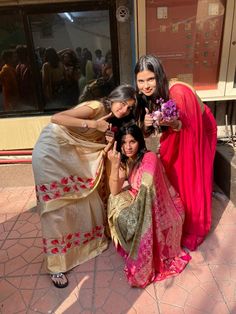 three women pose for the camera in front of a building with many people around them