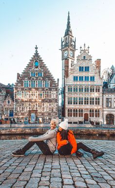 a man and woman sitting on the ground in front of buildings with a clock tower