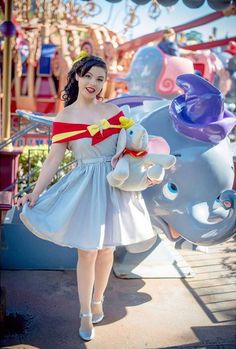 a woman in a dress standing next to an elephant at a amusement park, holding a stuffed animal