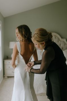 two women standing in front of a bed looking at the back of a wedding dress