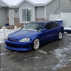 a blue car is parked in front of a house with snow on the ground and fence