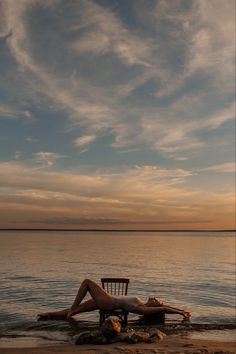 a woman laying on top of a wooden chair next to the ocean under a cloudy sky