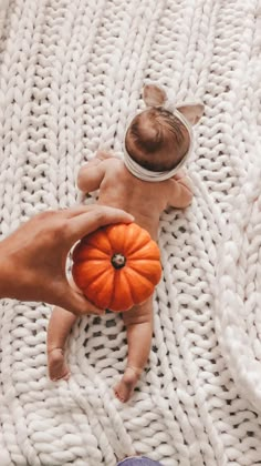 a baby laying on top of a white blanket next to a person holding a pumpkin