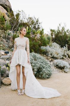 a woman in a wedding dress standing on a dirt road with bushes and rocks behind her