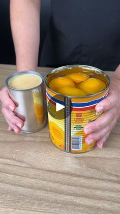 a woman holding two cans of oranges on top of a wooden table next to a cup of coffee