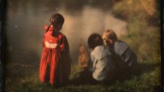 three children are sitting on the grass near a body of water, and one child is wearing a red dress