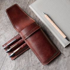 three pens and a notebook on a wooden table next to a pen holder with a notepad