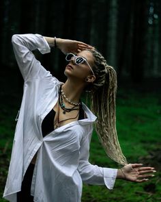 a woman with dreadlocks is standing in the woods wearing sunglasses and a white shirt