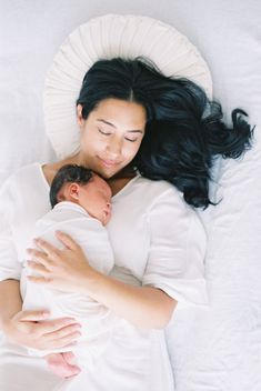 a woman holding a baby in her arms while laying on top of a white bed