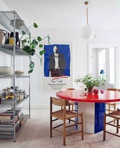 a red table and chairs in a white room with shelves filled with books, plants and other items