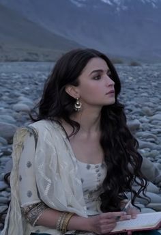 a woman sitting on rocks holding a book in her hands and looking off into the distance