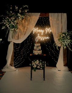 a wedding cake sitting on top of a table under a neon sign that says marry me