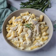 a white bowl filled with pasta covered in chicken and sauce next to a sprig of rosemary