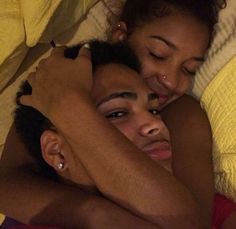 a man and woman laying in bed with their arms wrapped around each other looking at the camera