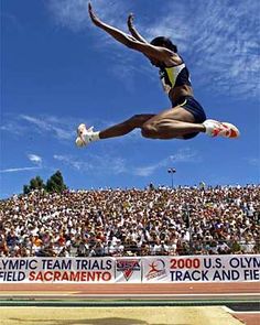 a woman jumping in the air on top of a track with an audience behind her