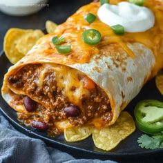 a black plate topped with a burrito covered in cheese and beans next to tortilla chips