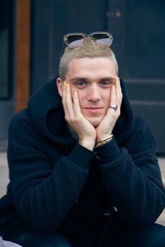 a young man wearing sunglasses sitting on the ground with his hands to his face and looking at the camera