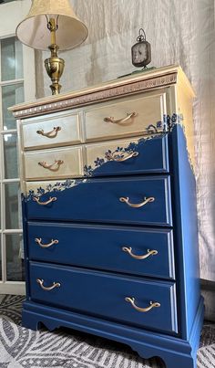 a blue dresser with gold handles in a room next to a lamp and curtains on the window sill