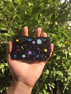 a hand holding up a small black box with planets and stars on it in front of some bushes