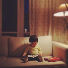 a little boy sitting on top of a couch reading a book next to a lamp