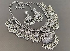 You can never go wrong with statement necklaces. Beautifully ornate with floral motifs , colourful stones & hanging pearls, this one of a kind necklace set will make you fall in love with it instantly. Handcrafted in german silver with oxidized silver plating , this glorious set has a breathtaking impact of instantly adding glam to your outfits. They look splendid with your sarees , low neck kurtas or even with your lehengas. Jewellery South Indian, Colourful Stones, Indian Choker Necklace, Antique Silver Necklace, Stone Necklace Set, Necklace Set Indian, Silver Jewellery Indian, Pearl Necklace Set, Studded Necklace