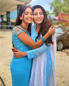 two young women hugging each other in front of a building with cars parked behind them